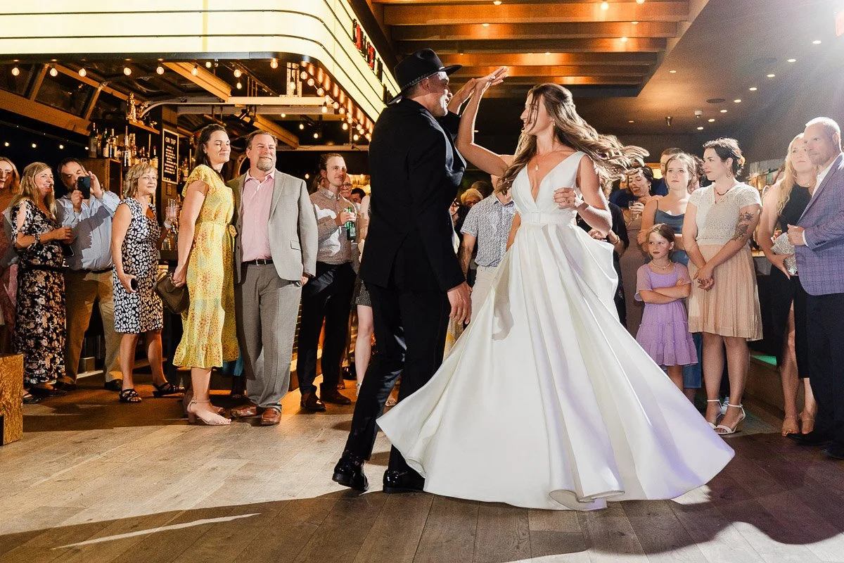 A bride in a flowing white gown and a man in a suit joyfully dance amid a lively crowd at a warmly lit indoor wedding reception.