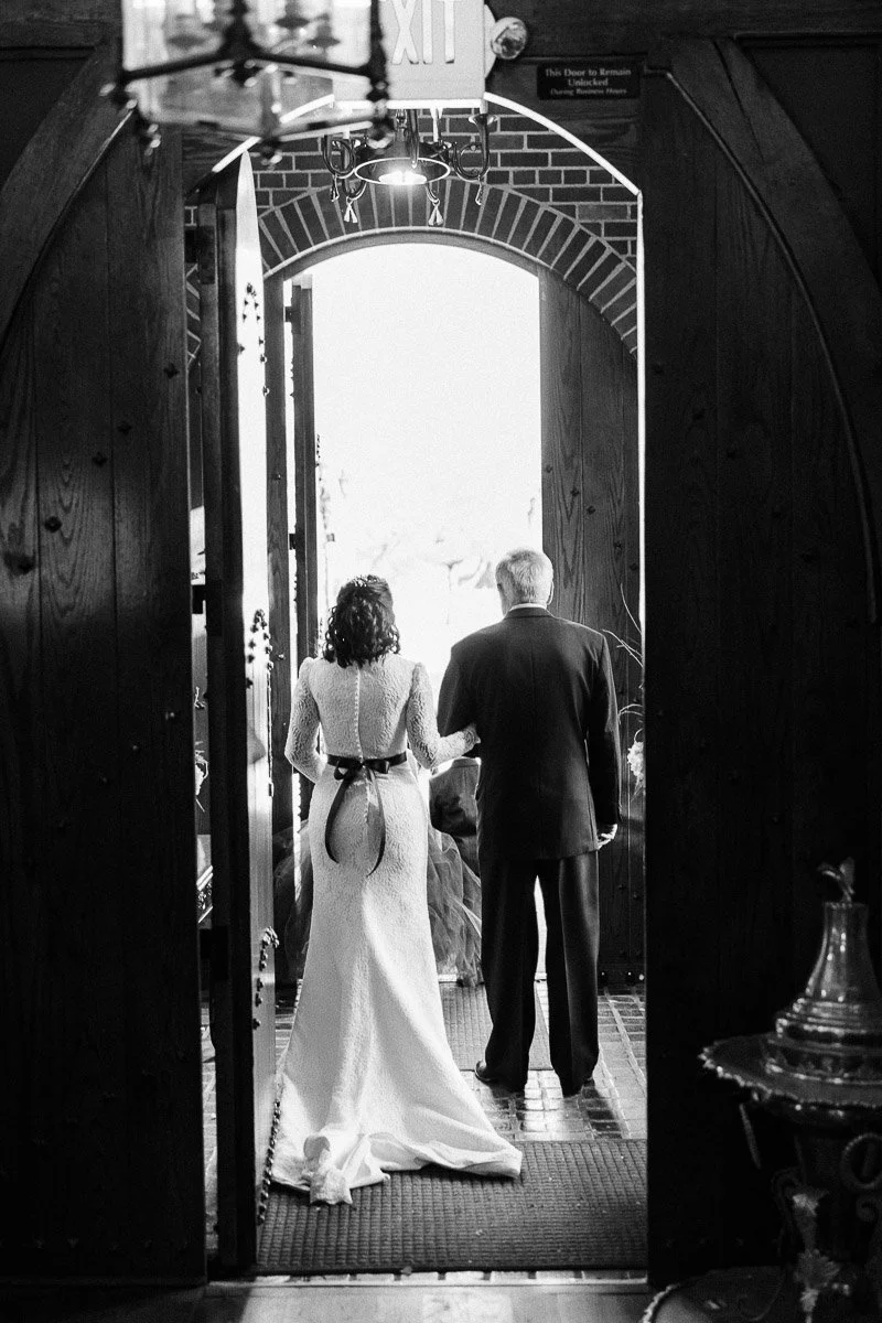 A bride in a lace gown and a man in a suit stand arm-in-arm, seen from behind, exiting through wooden doors into bright light. A chandelier hangs above.