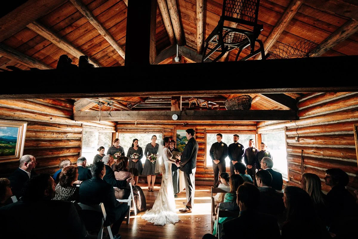 A couple stands at the altar in a rustic, sunlit cabin Ski Tip Lodge wedding, surrounded by their bridal party in dark attire and seated guests, creating an intimate, joyful ambiance.