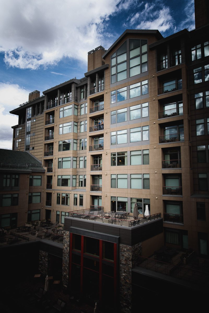 Tall modern Beaver Creek Westin building with glass windows under a partly cloudy blue sky. A wedding dress hangs in a door way. Calm atmosphere.