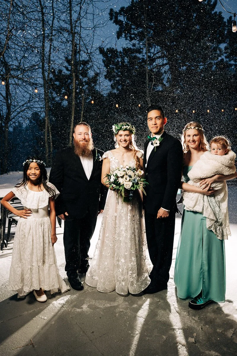A wedding party stands outdoors in falling snow. The bride and groom are in the center, both smiling. The group includes a child and a woman holding a baby.