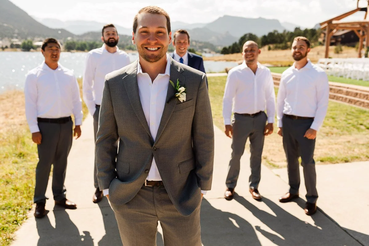 Groom in a gray suit stands smiling with groomsmen in white shirts behind him. Outdoor setting with mountains and a lake, clear blue sky.