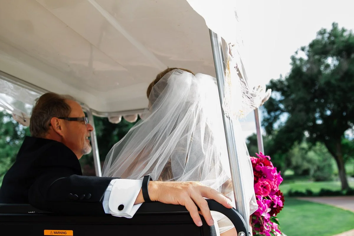 A bride in a white veil and a man in a suit ride a golf cart adorned with pink flowers, set against a backdrop of greenery and trees.
