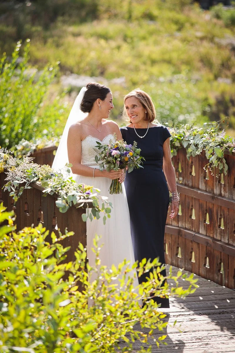 A bride in a white dress holds a bouquet, walking with an older woman in a navy gown on a flower-adorned bridge. They share a joyful, loving glance.