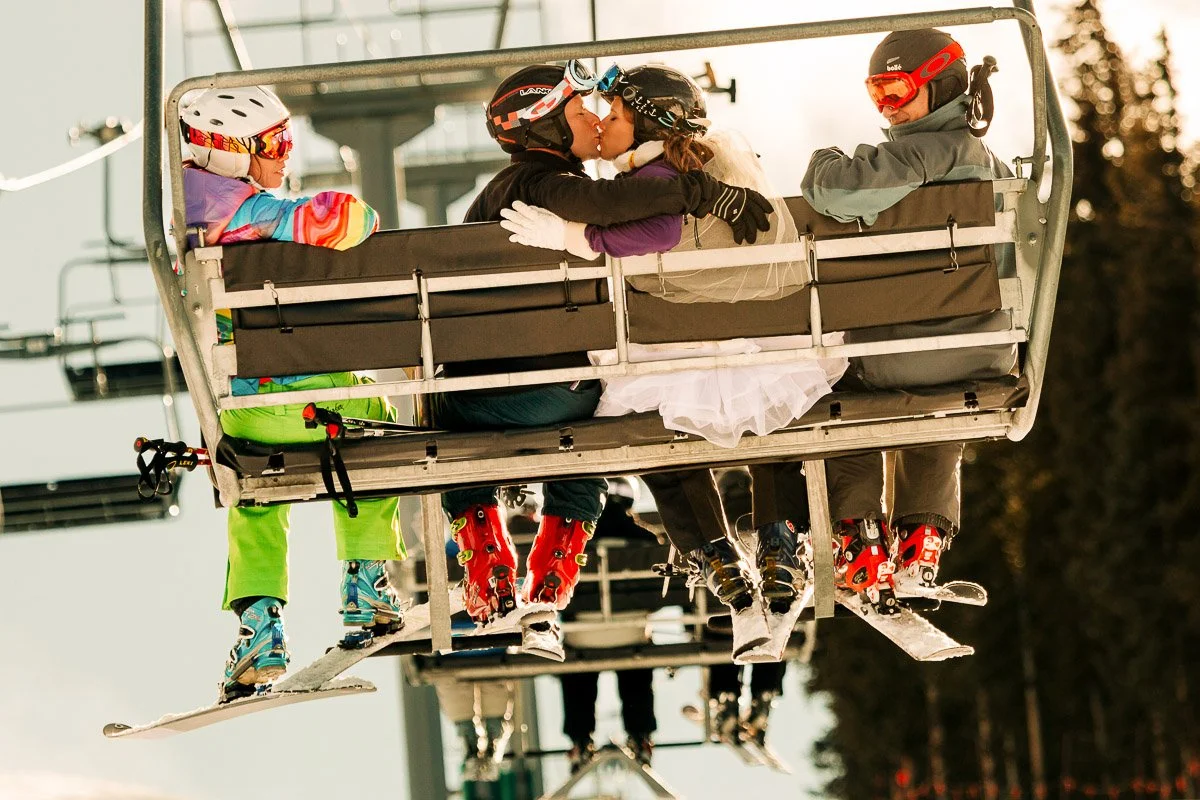 A couple in ski gear shares a kiss on a chairlift, with wedding attire visible. Two other skiers sit nearby. The scene is joyful and lively after a Park Hyatt wedding in Beaver Creek, Colorado.