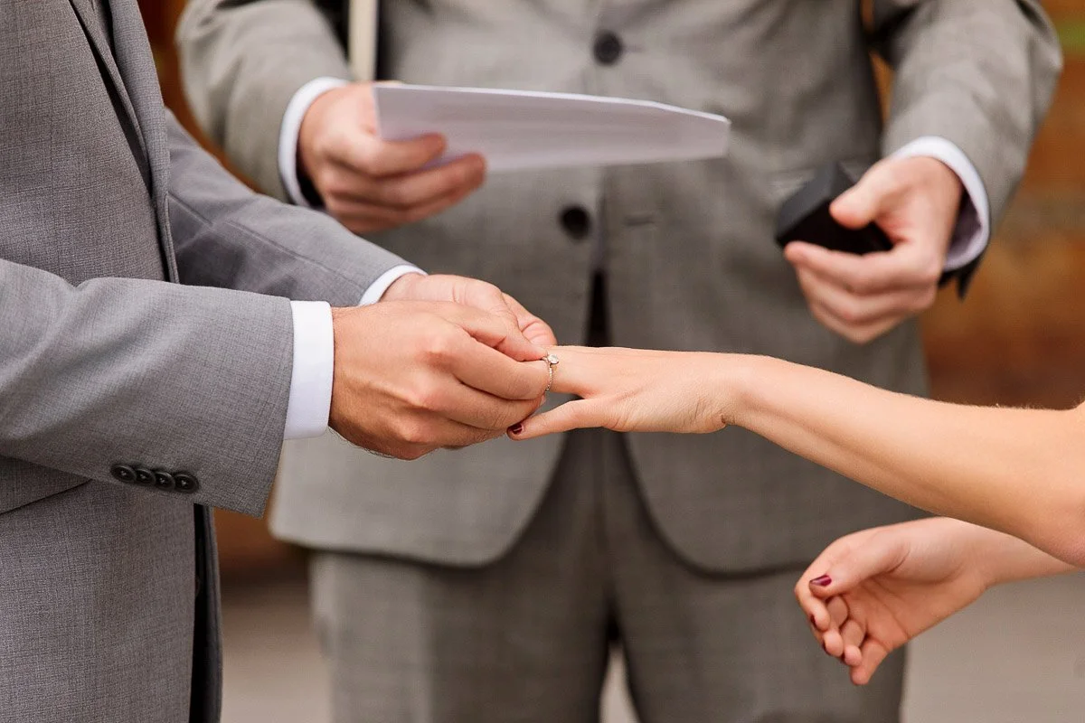 Groom in a gray suit placing a ring on bride's finger during wedding ceremony, officiant holding papers in the background, creating a formal, intimate atmosphere.