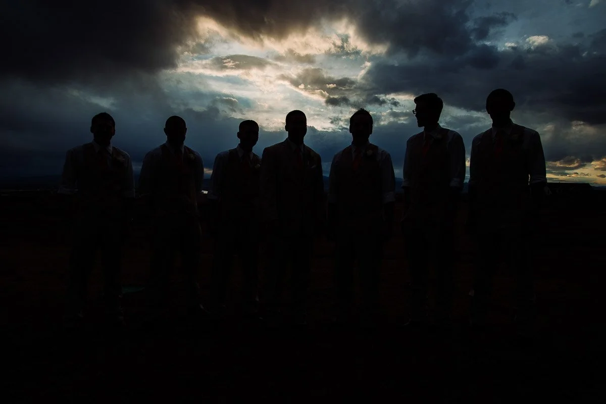 Silhouetted groomsmen stand against a dramatic sunset sky filled with dark clouds and soft light, creating a moody, tranquil atmosphere.