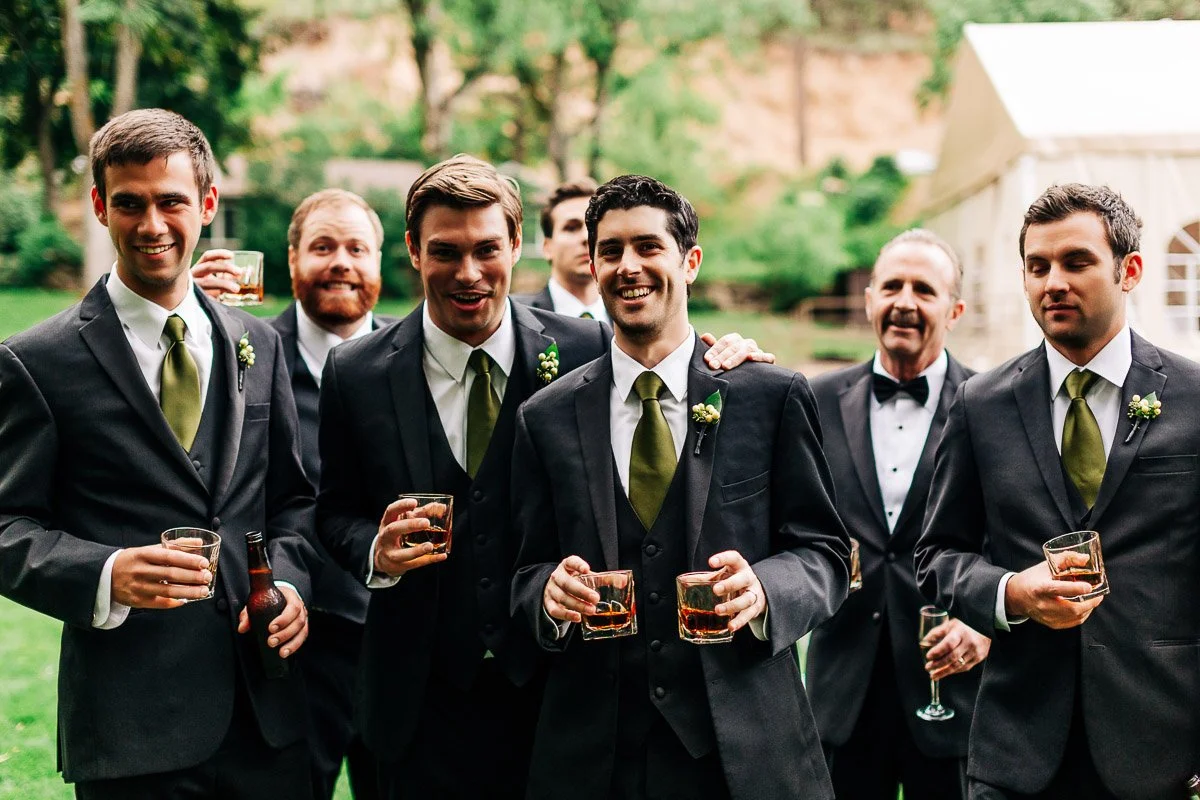 A group of men in matching black suits and green ties, smiling and holding drinks outdoors. The setting is festive and grassy with trees in the background.