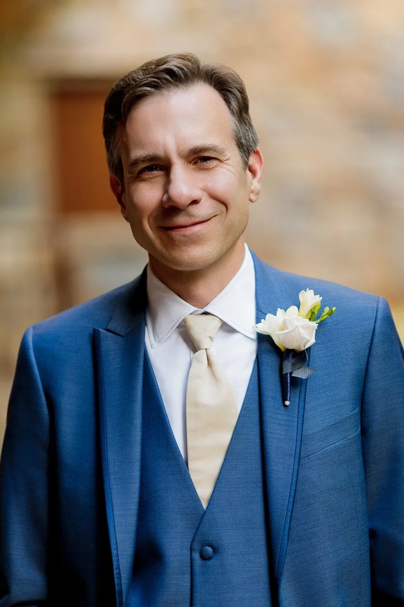 Smiling man in a blue suit with a beige tie and white boutonnière stands against a soft-focus background. Warm, cheerful expression.