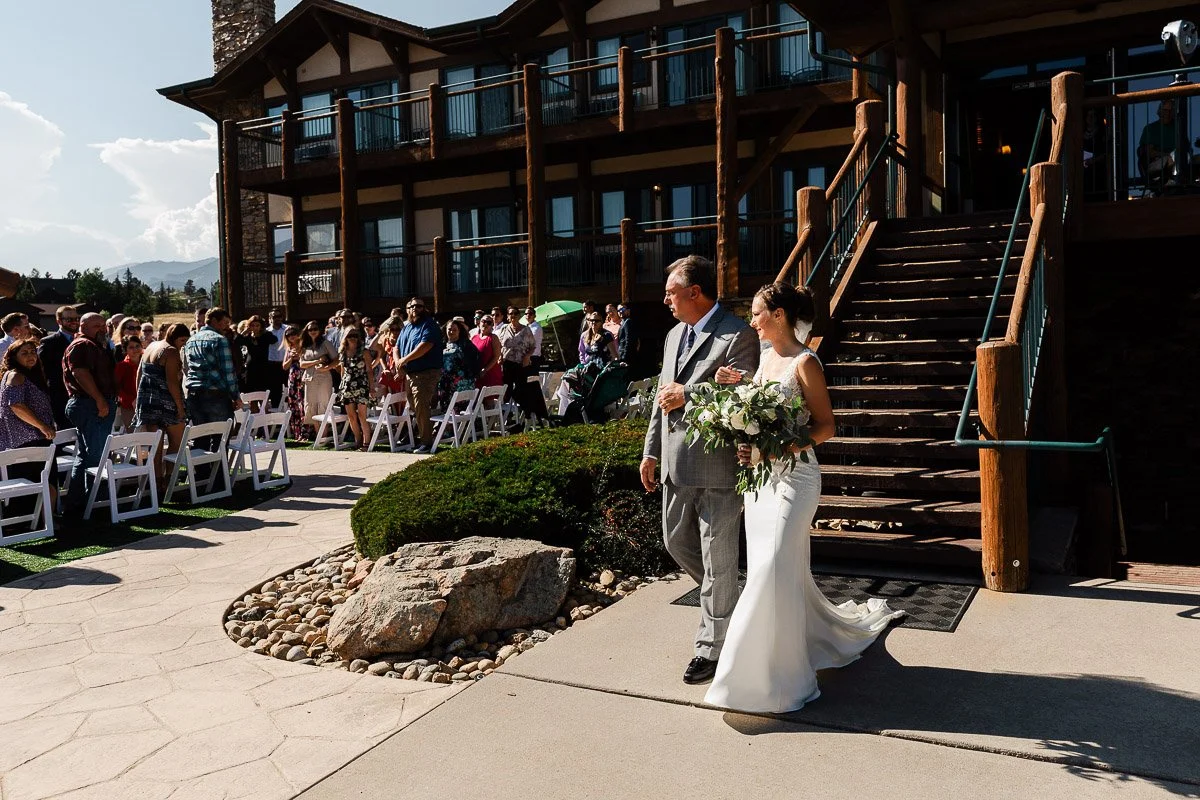 A bride in a white dress walks down outdoor stairs with an older man at a sunny wedding. Guests seated beside lawn watch, a rustic building in the background.