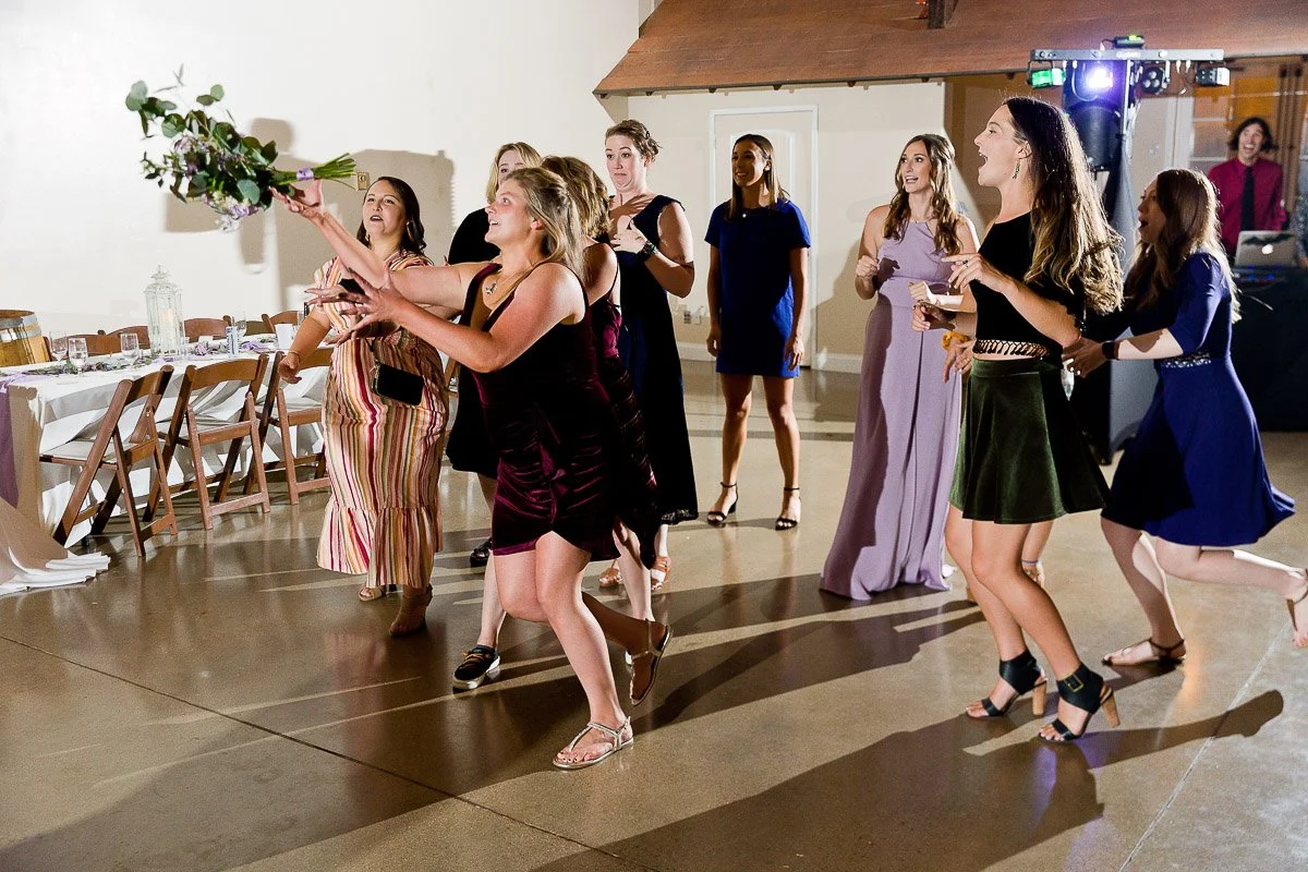 A group of women joyfully reach to catch a bouquet at a wedding reception. The scene is lively, with excitement and smiles amid elegant attire.