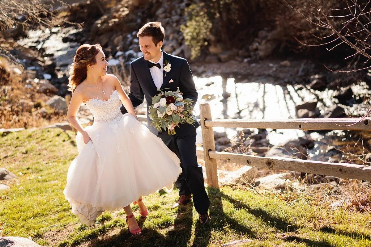 Bride in white gown and groom in dark suit joyfully walk near a sunlit stream and rustic fence, holding a bouquet. The scene conveys happiness and love.