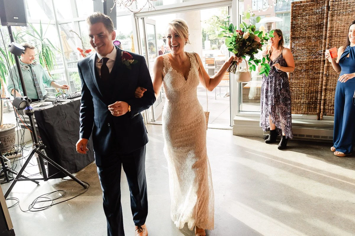 A joyful couple enters a sunlit venue, the woman holding a bouquet and wearing a white lace dress, with guests and a DJ celebrating nearby during a Coohills Restaurant wedding in Denver, Colorado.