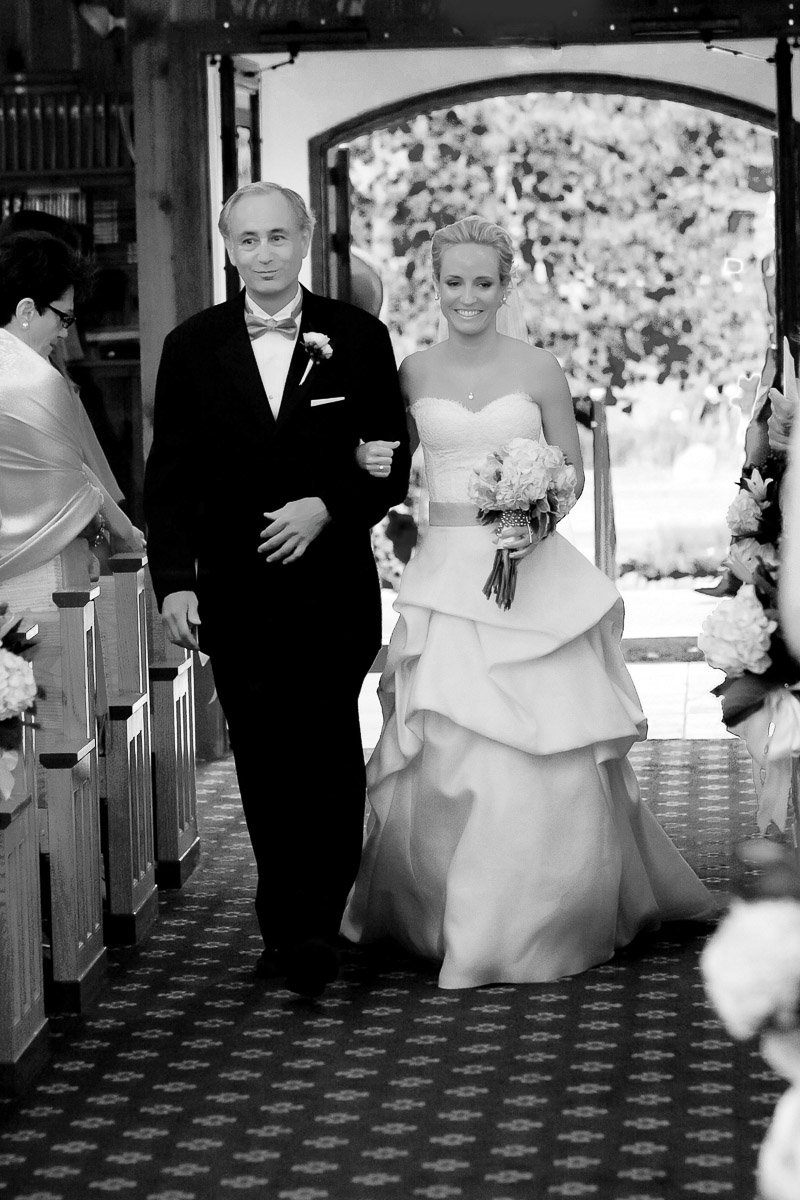 A smiling bride in a strapless gown and holding a bouquet walks down a flower-lined aisle with an older man in a tuxedo. The atmosphere is joyful and elegant.