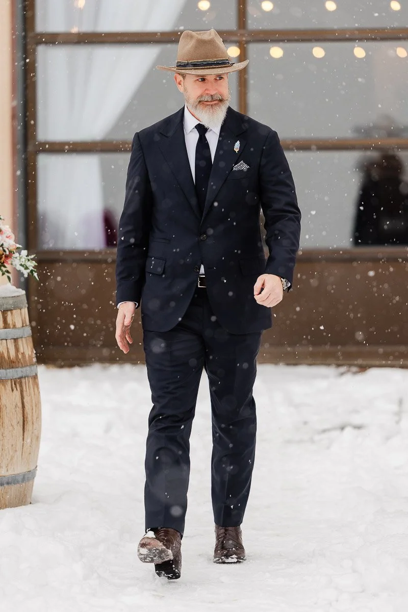 A man in a dark suit and tan fedora walks through snow, exuding confidence. Snowflakes fall around him, with warm lights visible in the background.