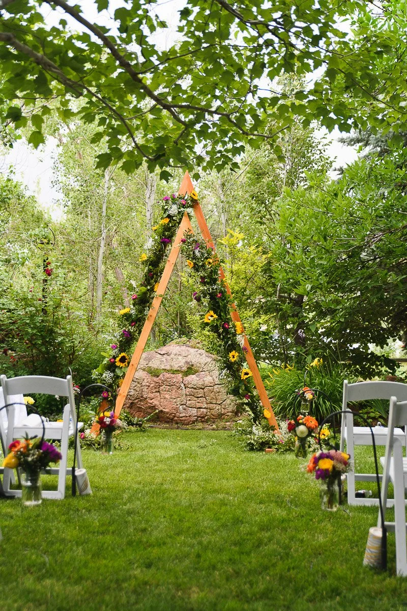 Triangular wooden wedding arch adorned with colorful flowers stands on a grassy area, surrounded by white chairs and vibrant bouquets, creating a serene outdoor setting.