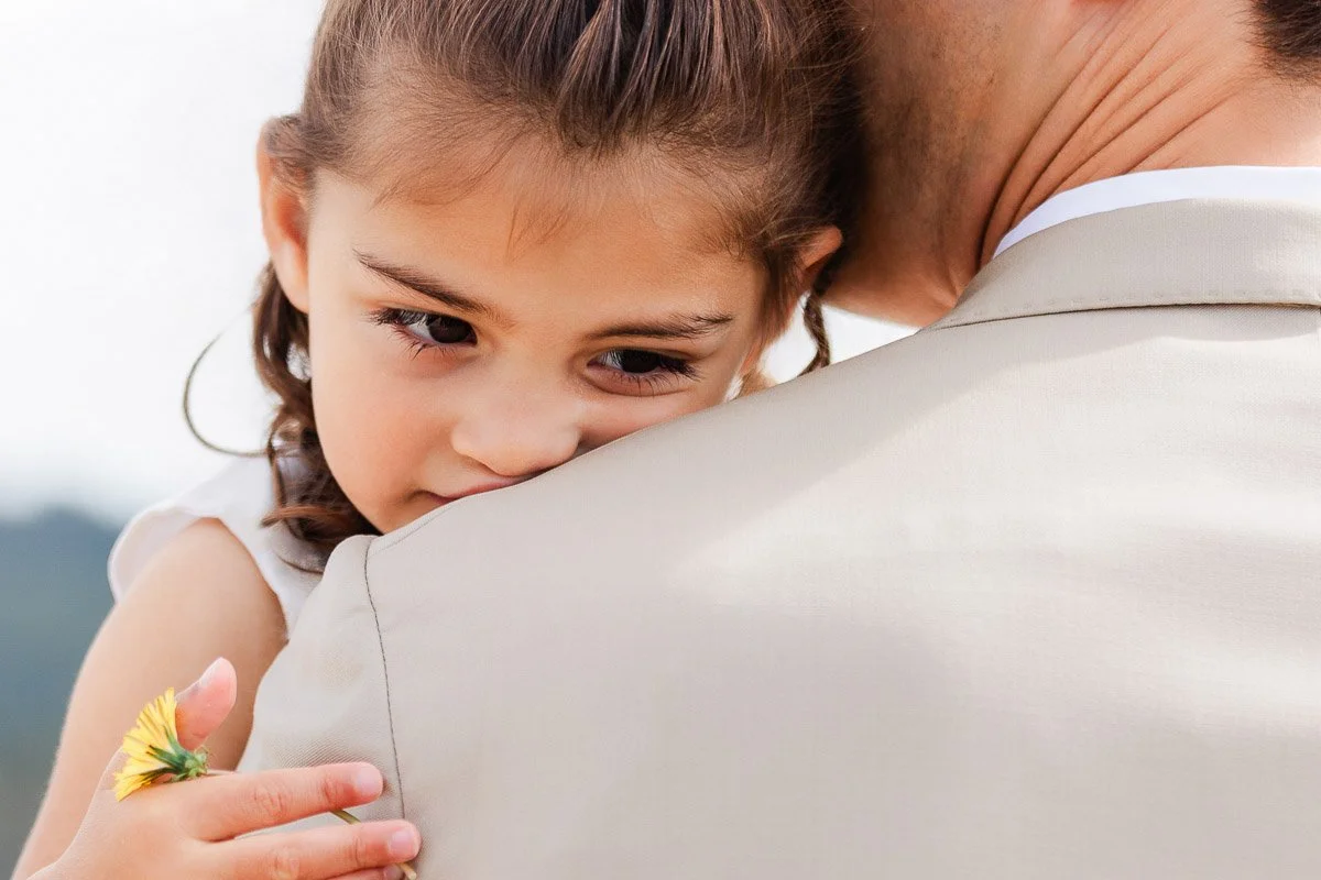 A young girl with a yellow flower in hand rests her head on an adult's shoulder, conveying comfort and affection. She appears pensive but serene.