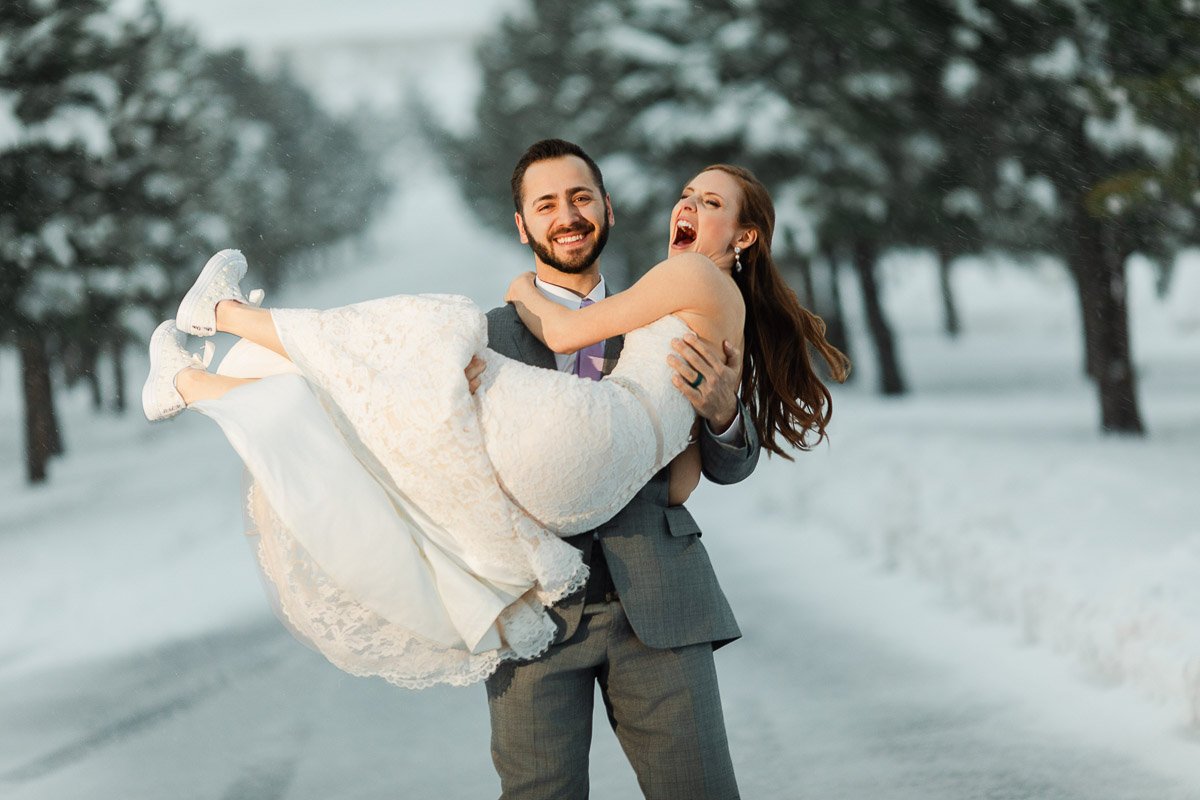A joyful couple in wedding attire stands on a snowy path lined with trees. The groom lifts the laughing bride, conveying happiness and warmth during a Spruce Mountain Wedding in Larkspur, Colorado
