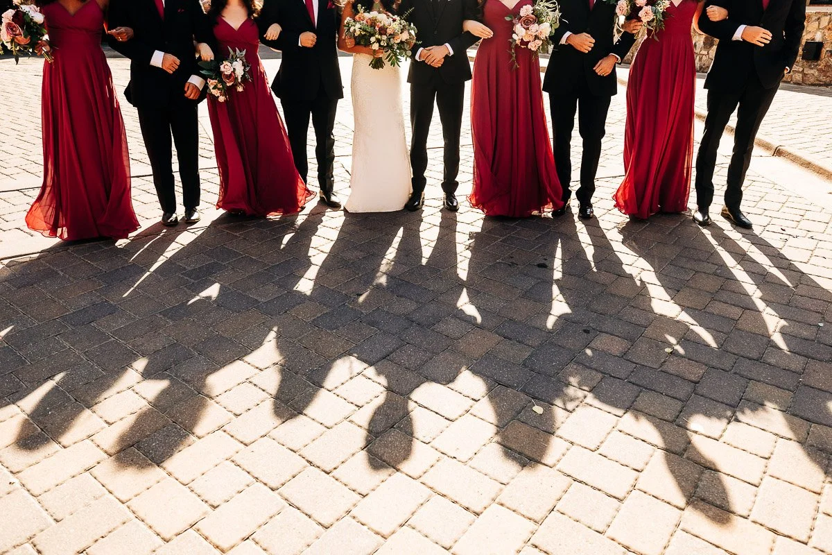 Wedding party stands on a sunlit cobblestone path, casting long shadows. Members wear burgundy dresses and black suits, holding floral bouquets.