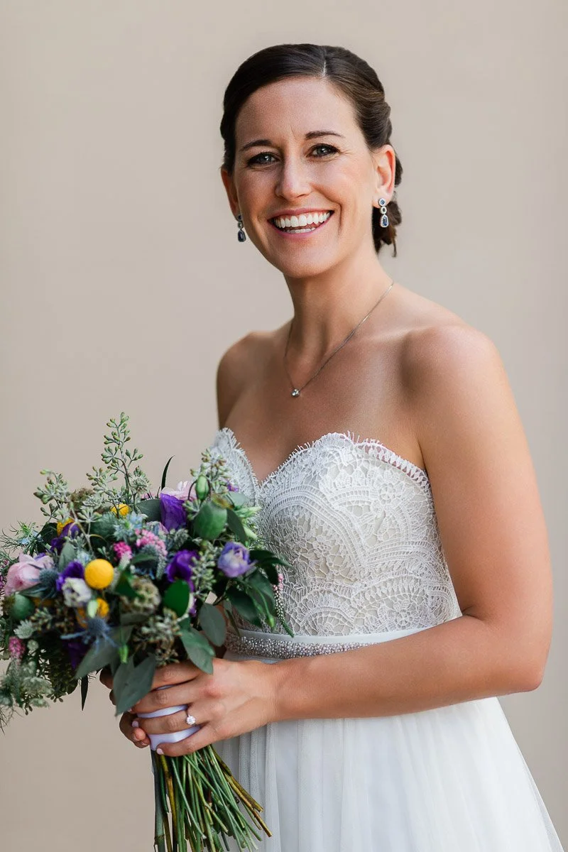 A smiling bride in a lace strapless wedding gown holds a colorful bouquet with purple, yellow, and pink flowers. The background is a neutral beige.