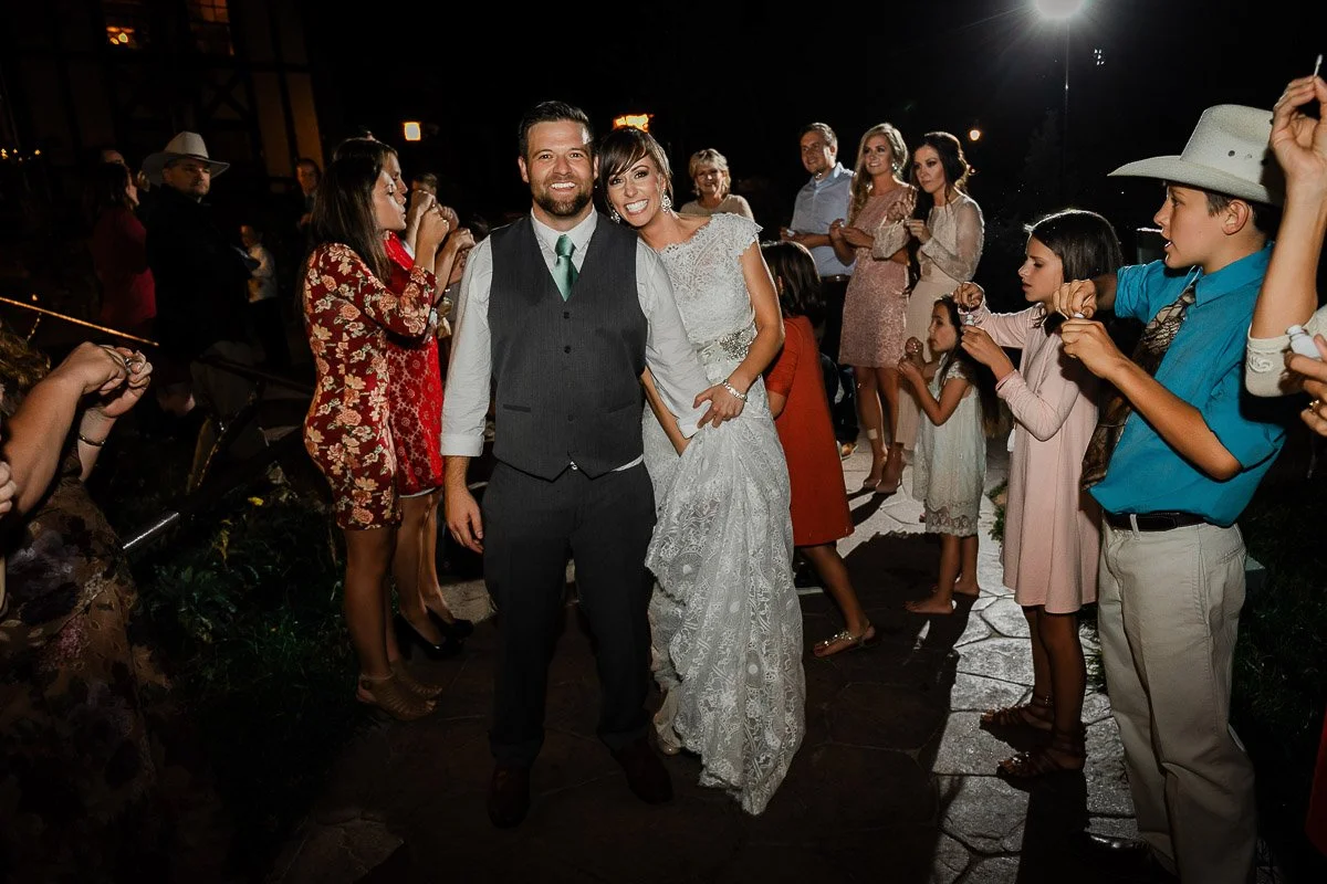 A joyful couple in wedding attire stands surrounded by cheering guests blowing bubbles at night. The scene is lively and festive, capturing celebration.