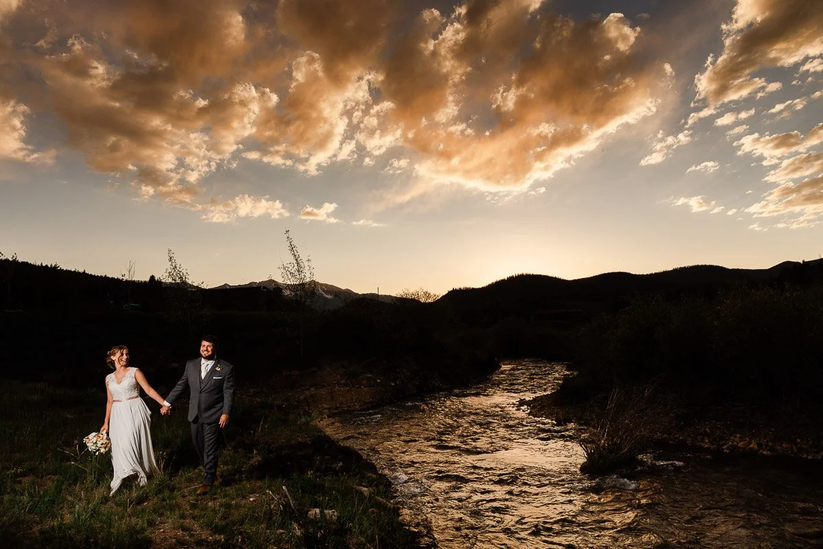 A couple, holding hands, walks along a stream at sunset. The sky glows with orange clouds, creating a romantic and serene atmosphere.