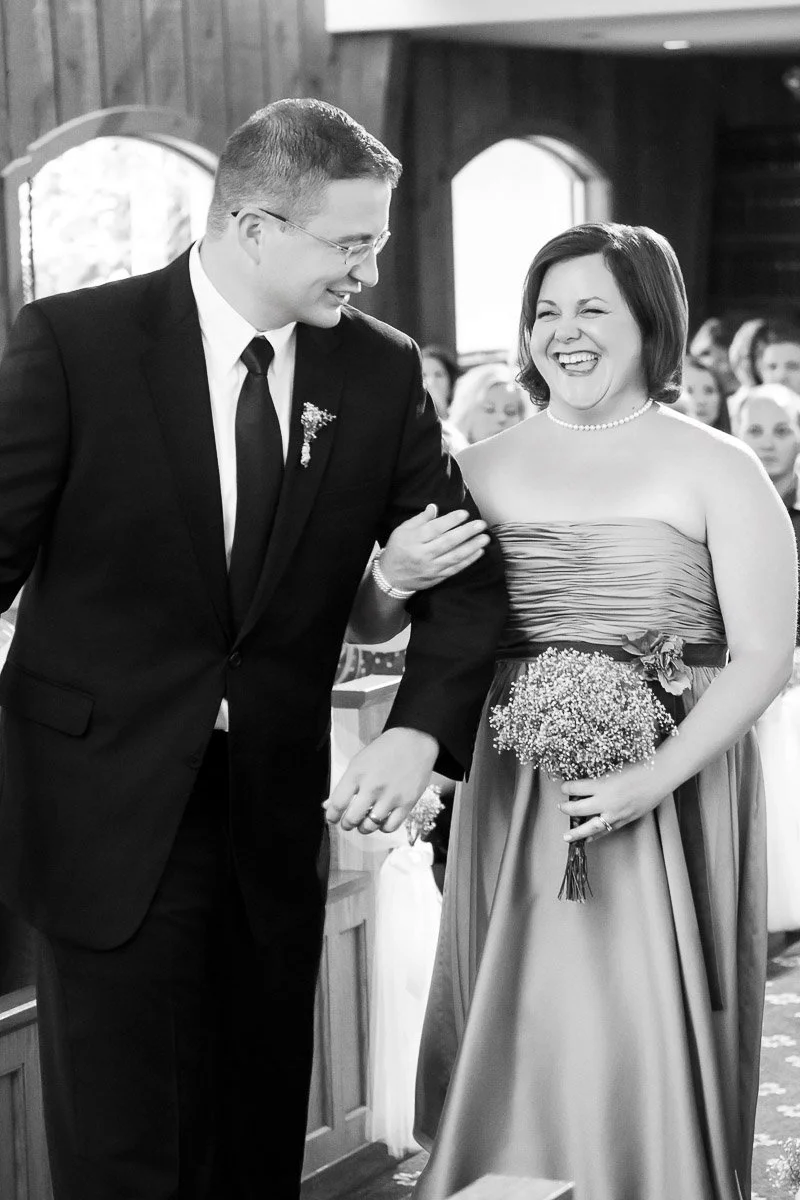 A joyful couple walks down the aisle in a church, smiling warmly. The man wears a suit, and the woman is in a strapless dress holding a bouquet.