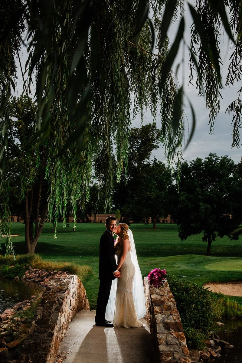 Bride and groom stand on a stone bridge under willow branches, holding hands. The lush green Lakwewood Country Club golf course and vibrant flowers create a romantic scene.