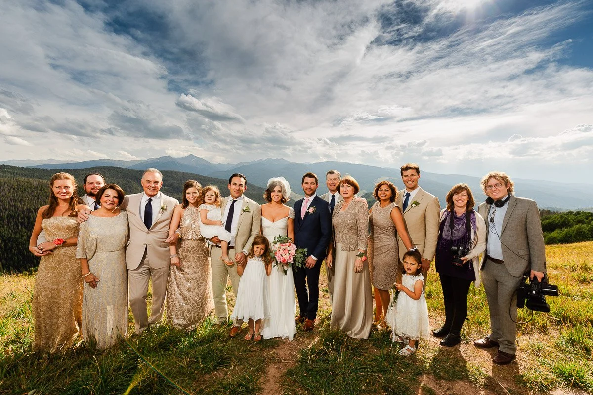 A joyful wedding party poses in formal attire on a sunny hillside, with a scenic mountain landscape in the background. Smiles convey happiness.