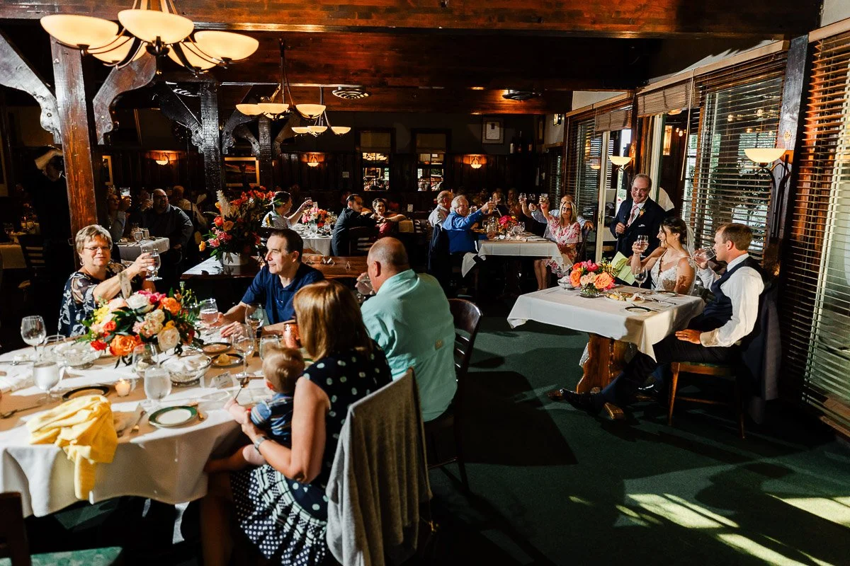 Guests seated in a warmly lit restaurant, toasting at a wedding reception. Floral centerpieces adorn tables, creating a cheerful and celebratory atmosphere.