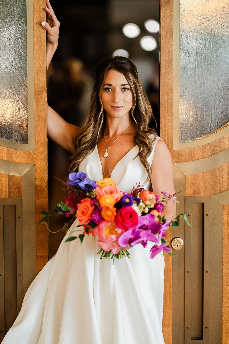 A woman stands in a Elizabeth Hotel doorway, wearing a white dress, holding a vibrant bouquet of pink, orange, and purple flowers. She has a calm, poised expression.
