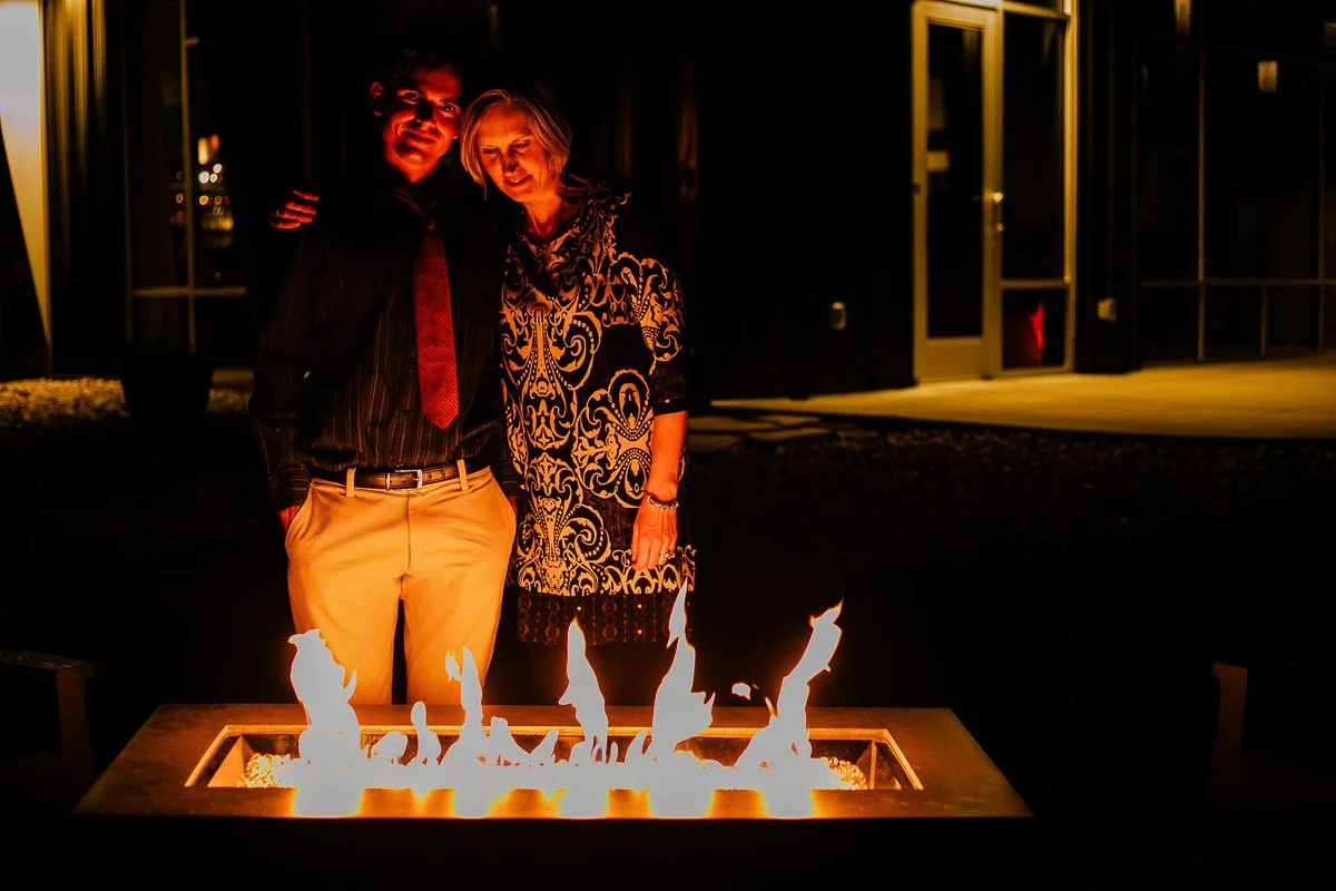A couple stands closely together, smiling warmly beside a glowing fire pit at night. The ambiance is cozy and intimate, with soft, flickering flames.