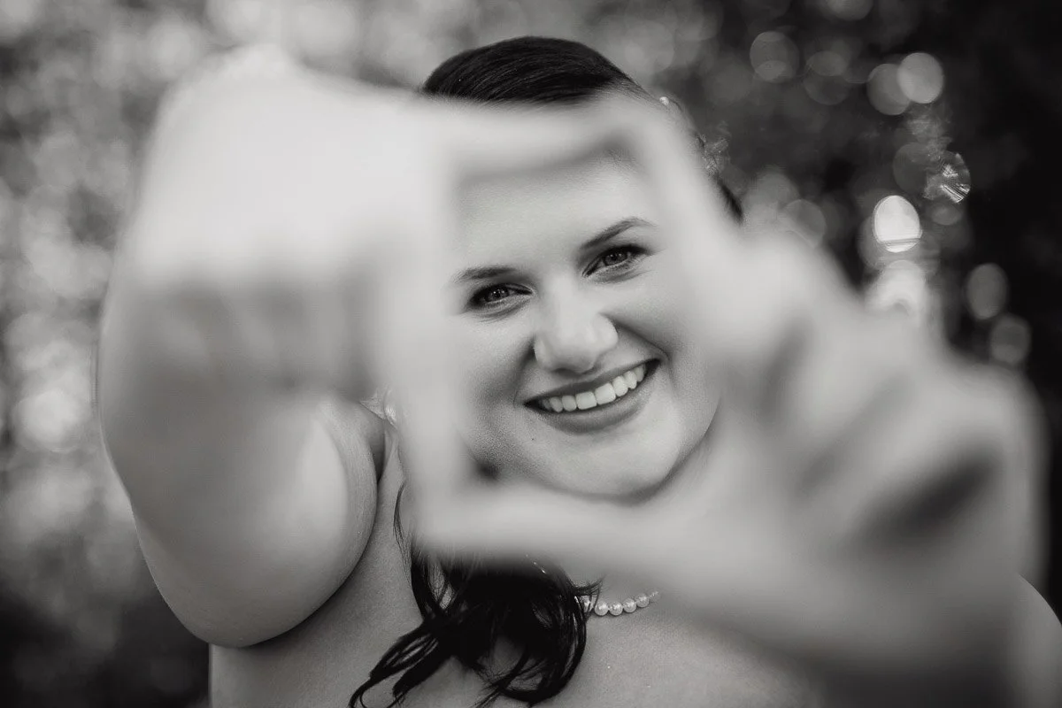 Black and white image of a woman smiling, framing her face with her hands. Soft focus background creates an intimate and joyful atmosphere.