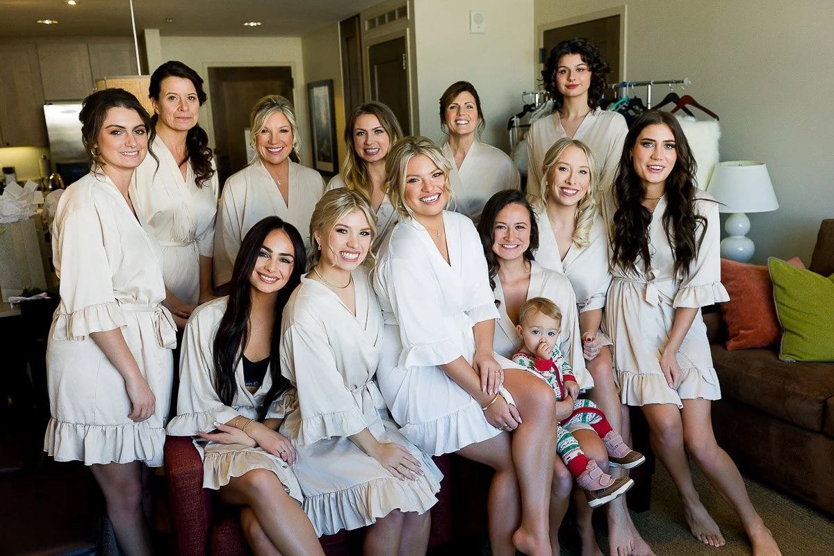 A group of women in matching cream robes and a toddler in pajamas smile warmly in a cozy living room, creating a joyful, relaxed atmosphere.