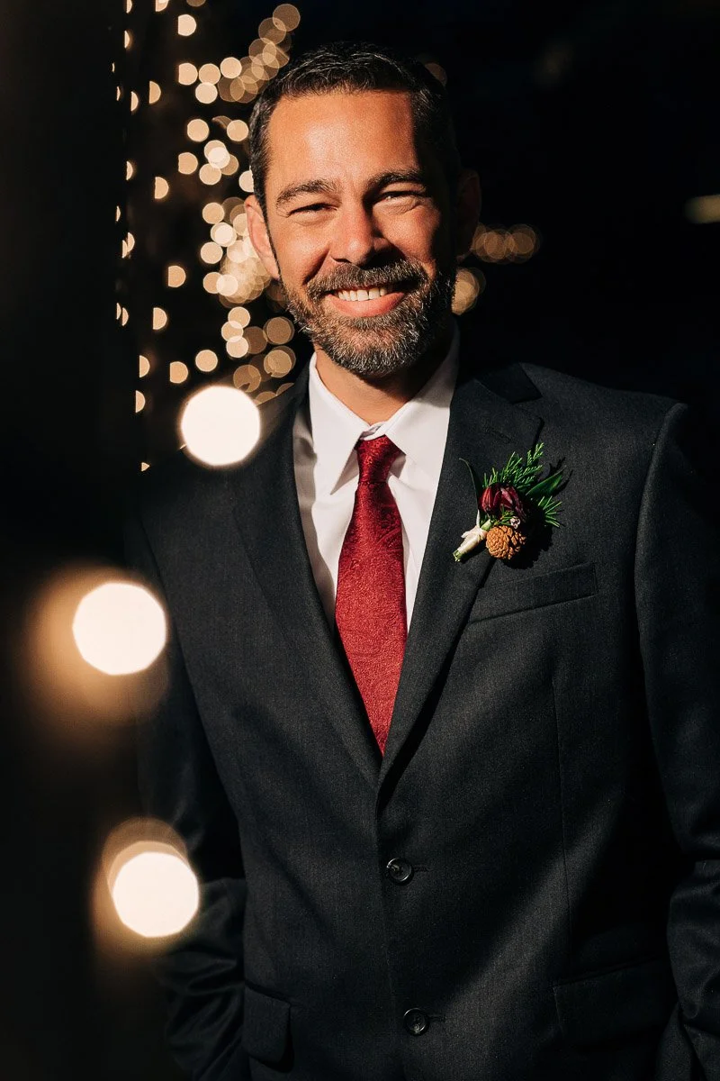 Smiling man in a dark suit with a red tie and boutonniere stands against a backdrop of blurred lights, creating a warm, festive ambiance.
