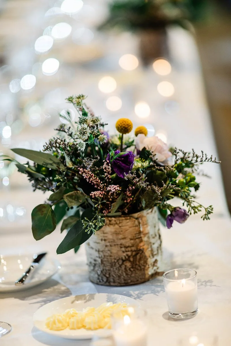 Elegant table setting with a rustic floral centerpiece in a birch vase. Surrounding candles and soft lighting create a warm, inviting atmosphere. Blurred background.