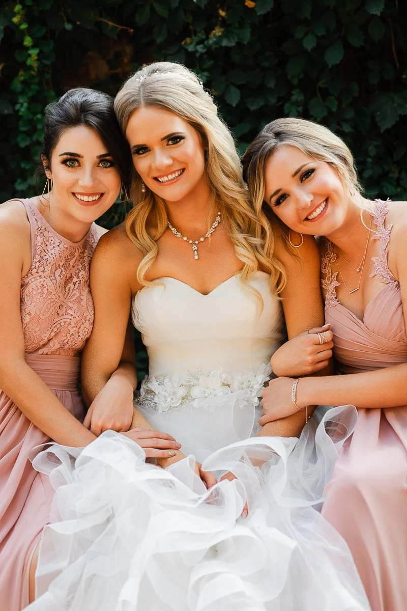Three women smiling warmly; the center one in a wedding dress, flanked by two in pink dresses, exuding joy and closeness, against a leafy backdrop.