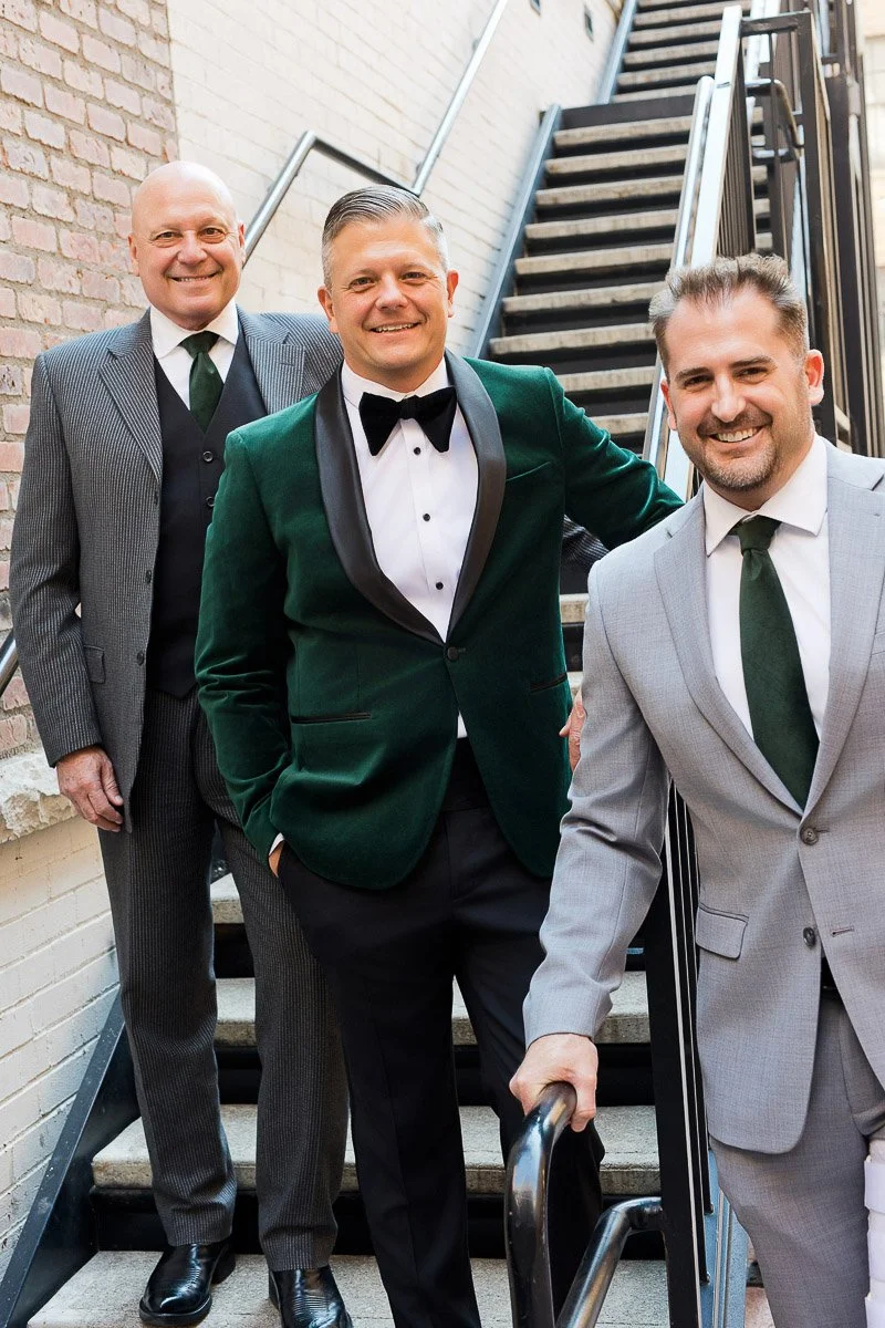 Three men in formal attire stand on outdoor stairs. One wears a green velvet jacket, another a striped suit, and the third a gray suit, all smiling warmly.