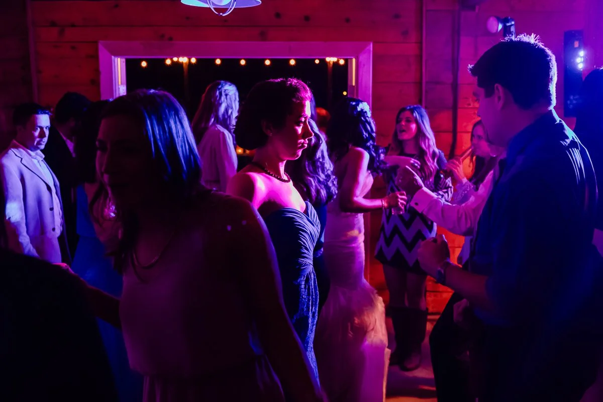 A lively party scene with people dancing under vivid pink and purple lighting. The room has wooden walls, adding a cozy atmosphere to the festivities.