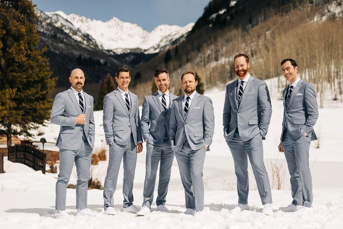 Six men in light gray suits and ties stand on snow, smiling against a backdrop of snow-capped mountains and trees, conveying a joyful, formal tone.