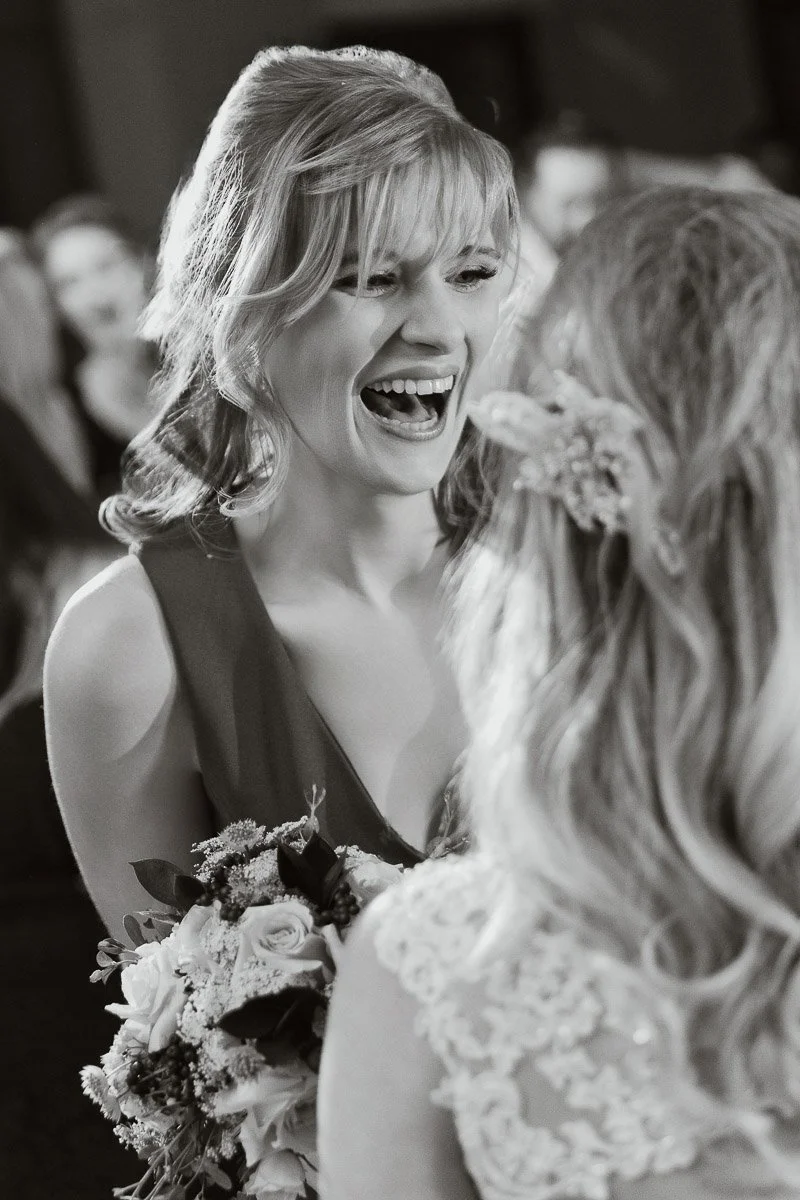 A woman laughing joyfully holds a bouquet and faces another woman with floral hair accessories in a lace dress, in a lively, festive setting.