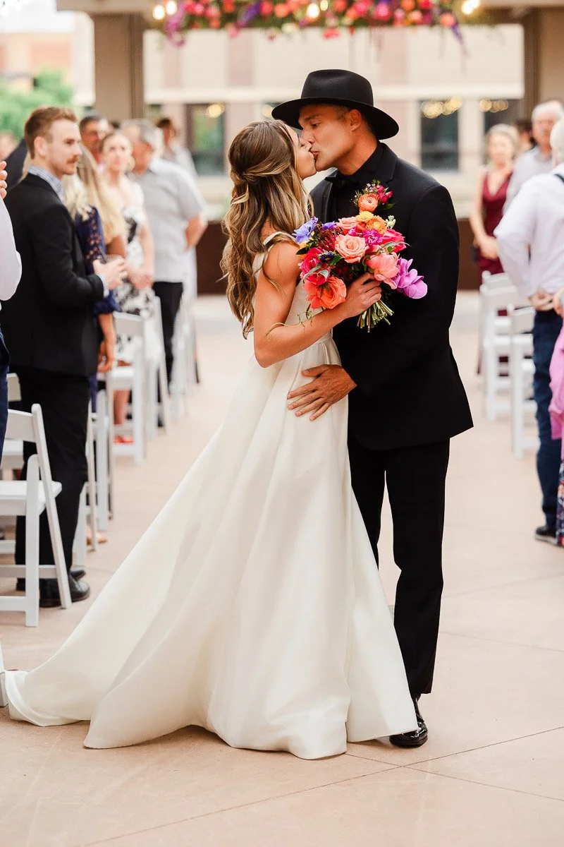 A bride in a flowing white gown kisses a groom in a black suit and hat. He holds vibrant, colorful flowers. The setting is joyful, with guests looking on.