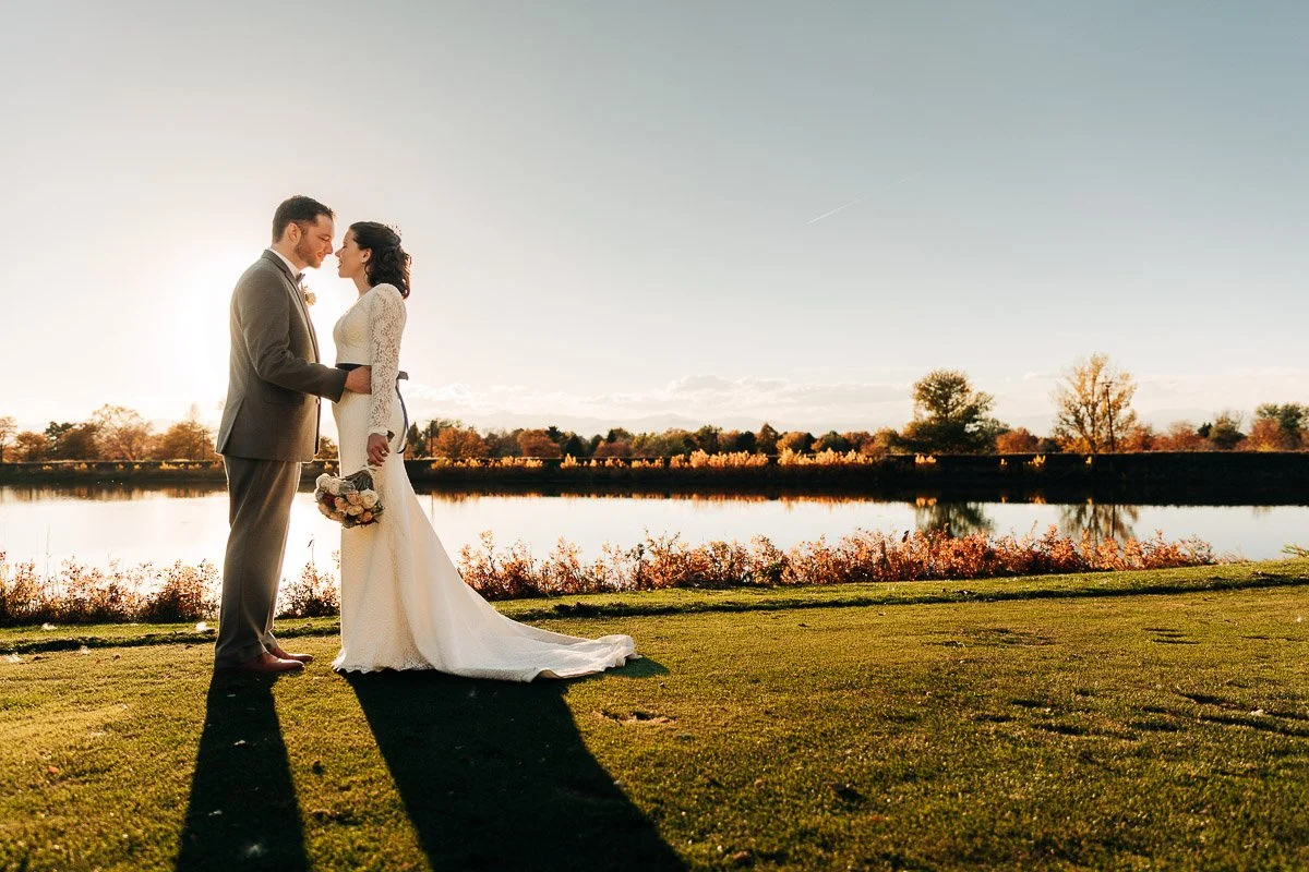 A bride and groom stand on grass by a serene lake at sunset, their silhouettes softly lit. The bride holds a bouquet. The scene conveys romance and tranquility.