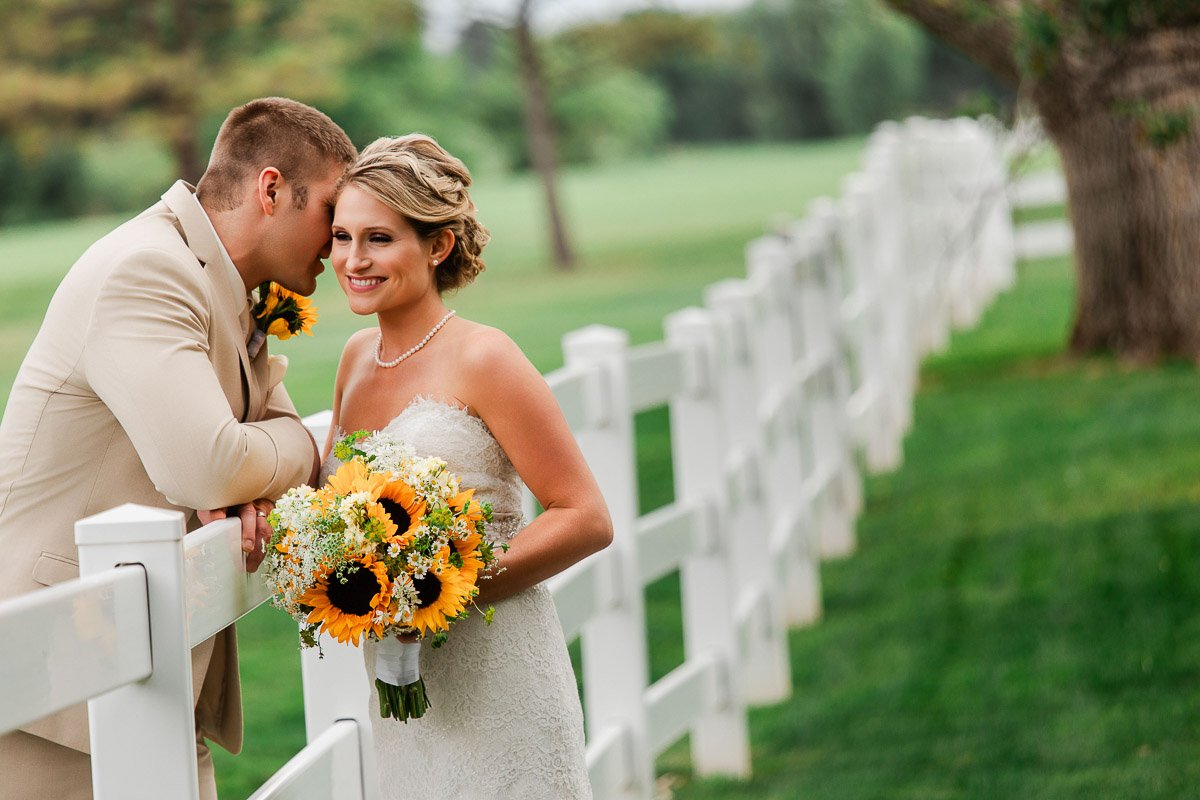 A bride in a strapless gown holds a sunflower bouquet, while smiling beside a groom in a beige suit. They stand near a white fence on a lush lawn.