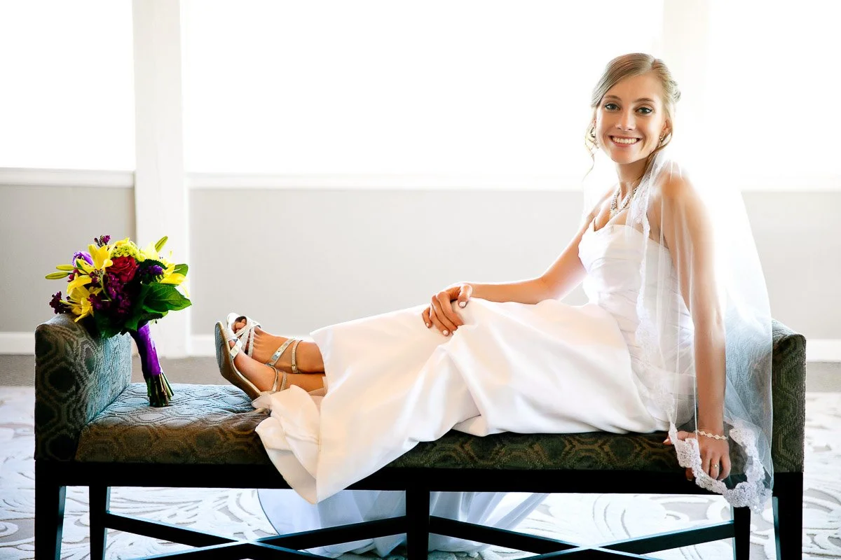 Bride in a white dress and veil smiling brightly, lying on a bench. Bright bouquet of purple and yellow flowers beside her. Bright, joyful atmosphere.