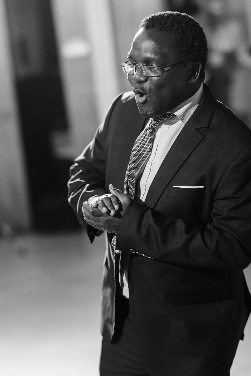 A man in a suit and tie speaks passionately, gesturing with his hands, in a softly lit room. The black-and-white image conveys a serious tone.