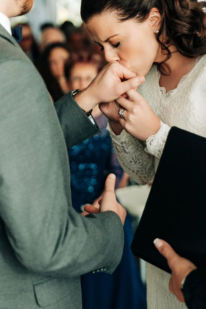 A bride in a lace dress kisses the groom’s hand during a wedding ceremony. The atmosphere is intimate and emotional, with guests blurred in the background.
