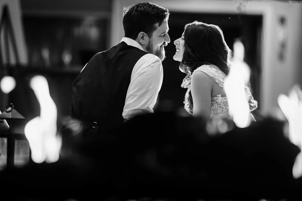 A couple, dressed formally, smiling and leaning in close amidst soft, out-of-focus flames in foreground. The setting is intimate and romantic.