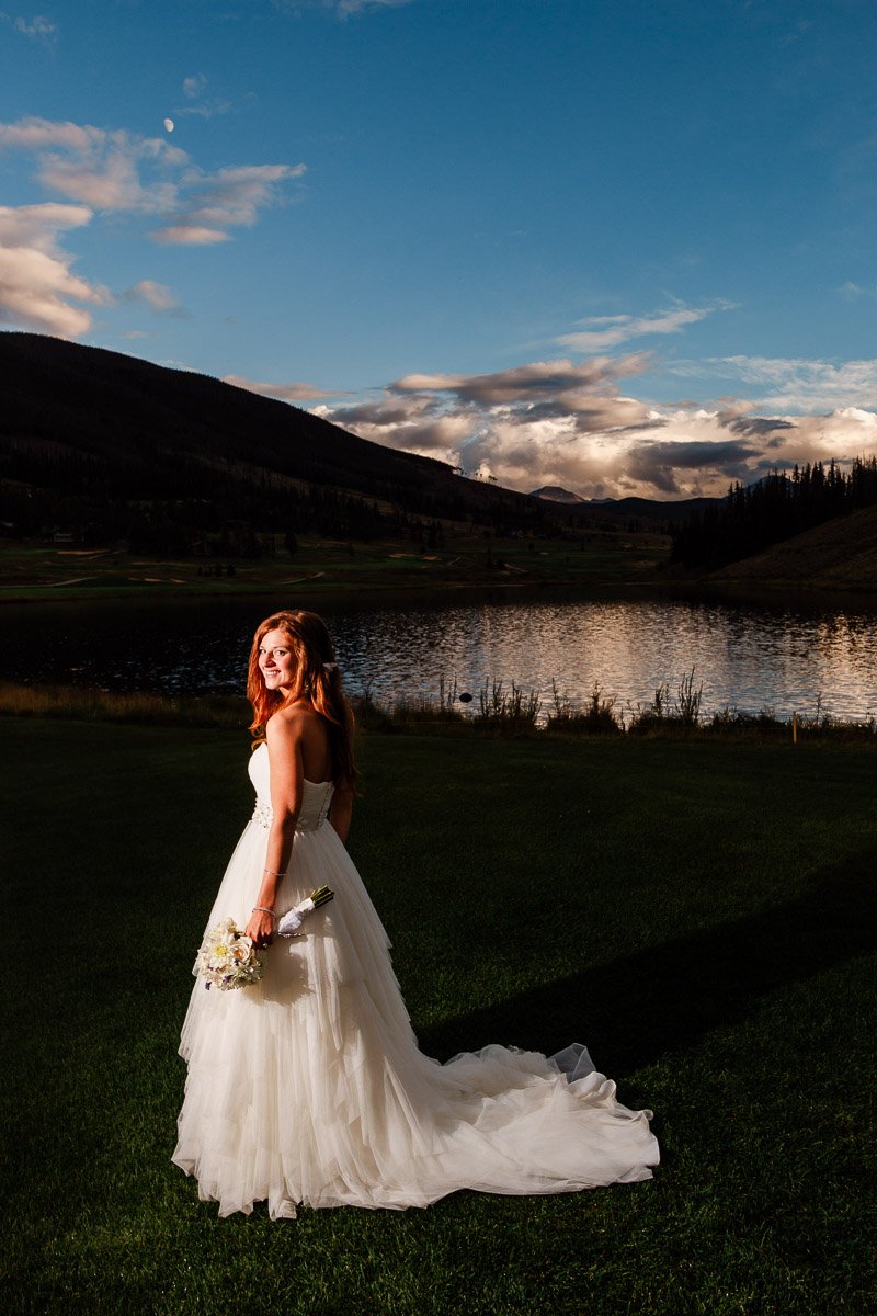 Bride in a flowing white gown stands on green grass by a serene lake at sunset, holding a bouquet, with mountains silhouetted in the background captured by keystone wedding photographer tomKphoto