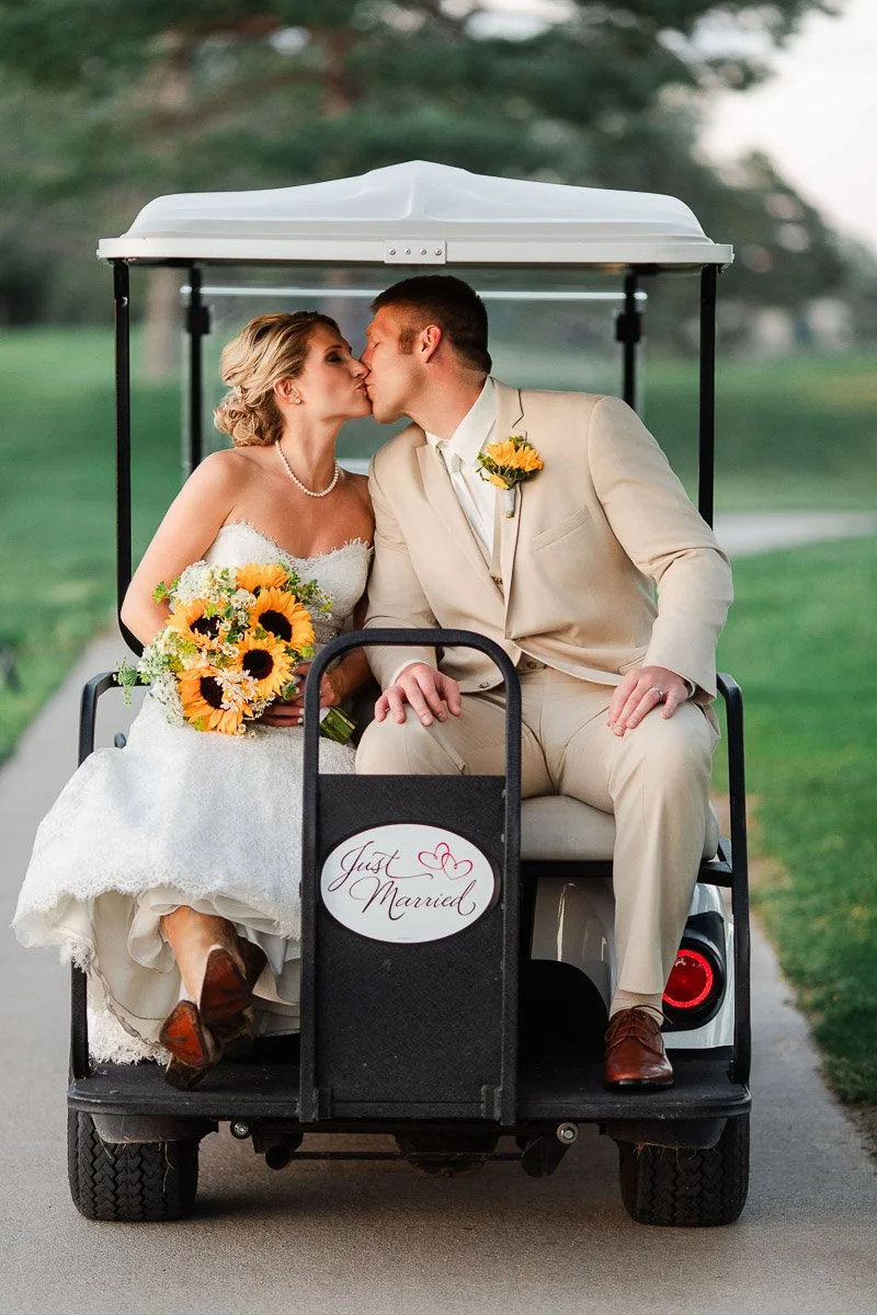 A newlywed couple kisses on a golf cart adorned with a "Just Married" sign. The bride holds sunflowers, conveying joy and celebration in a scenic outdoor setting.